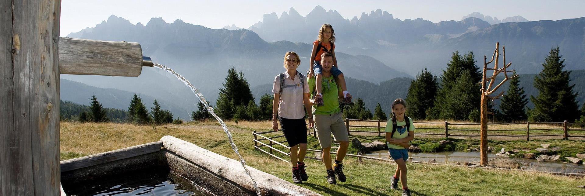 Eine Familie wandert an einem Brunnen in den Dolomiten vorbei, im Hintergrund die imposanten Gipfel der Geislergruppe.