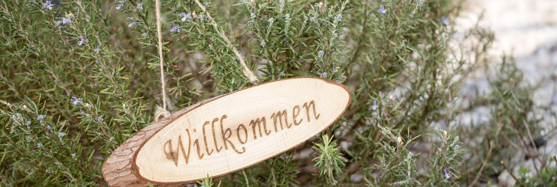 Ovales Holzschild mit der Aufschrift „Willkommen“, aufgehängt an einem blühenden Rosmarinstrauch.