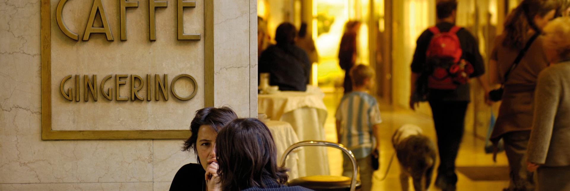 Menschen im Gespräch vor dem Caffè Gingerino in Bozen, im Hintergrund Passanten unter den Lauben.