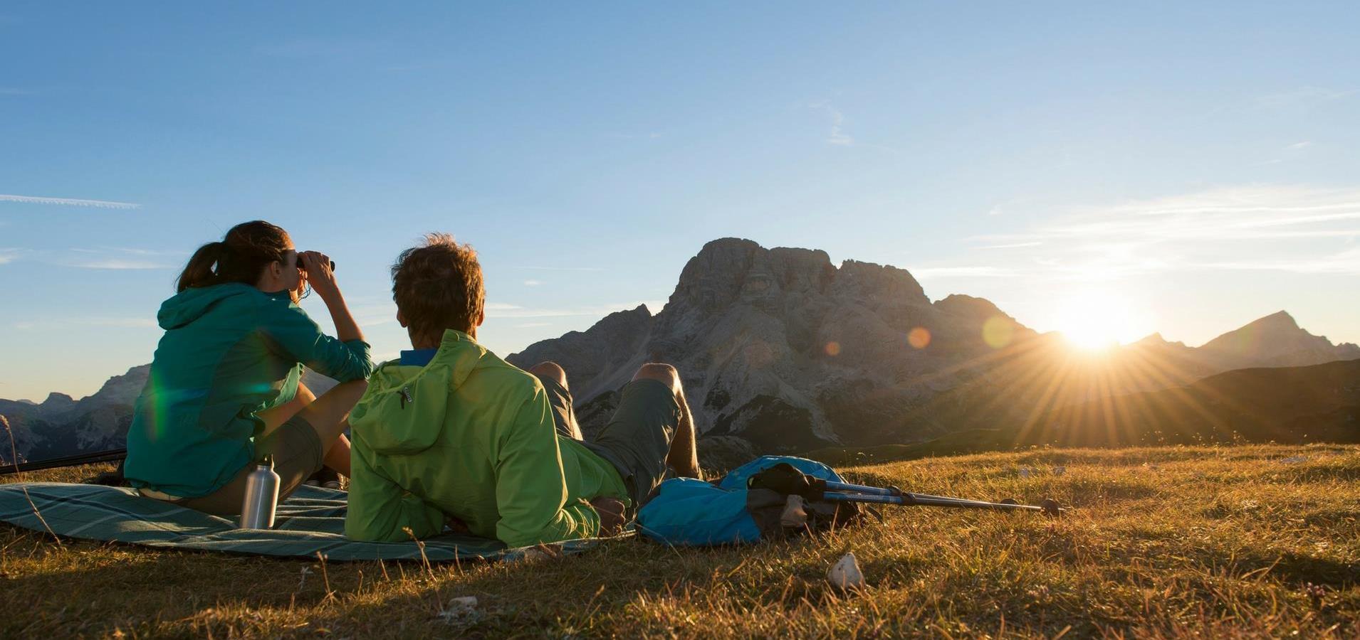 Two hikers sitting on a blanket watching the sunset over the mountains.
