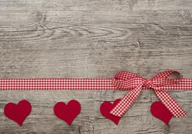 Red checkered ribbon with bow and four red felt hearts on a rustic wooden background.