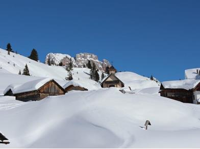 Villaggio alpino innevato con baite in legno e cappella, circondato da montagne sotto cielo limpido.