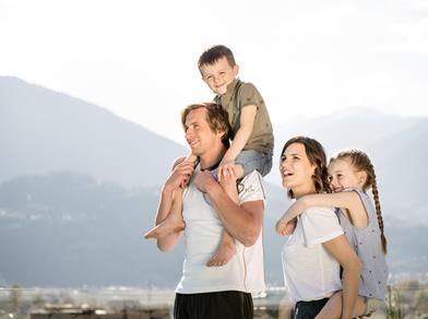 Eine fröhliche Familie mit zwei Kindern genießt den Ausblick auf die Berge – die Eltern tragen die Kinder auf den Schultern.