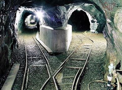 Galleria illuminata di una miniera con binari che si diramano in due direzioni. Le pareti sono di roccia grezza e sul lato sinistro si trova un simbolo con martello e mazza incrociati.