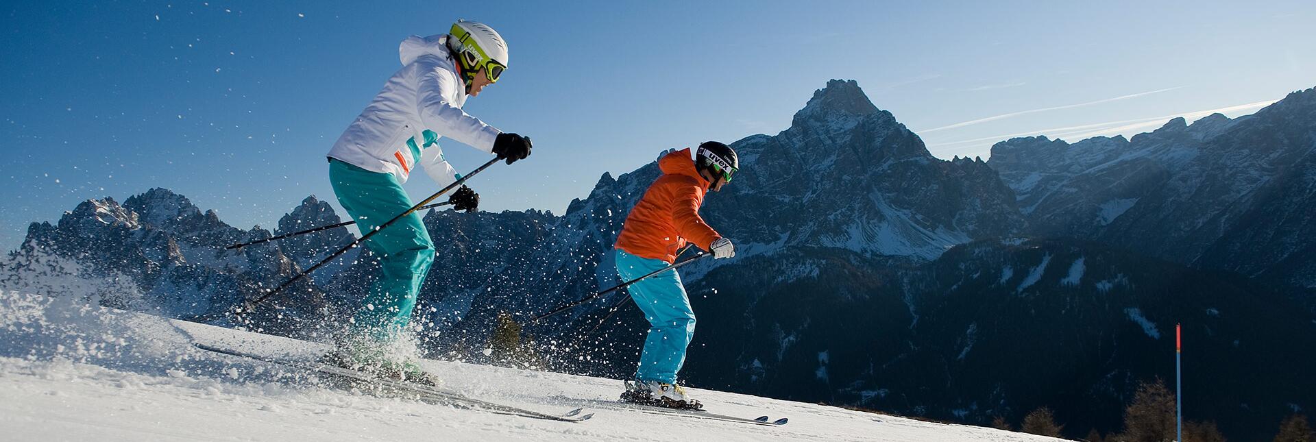 Two skiers descend a slope surrounded by snowy mountains and a clear blue sky.
