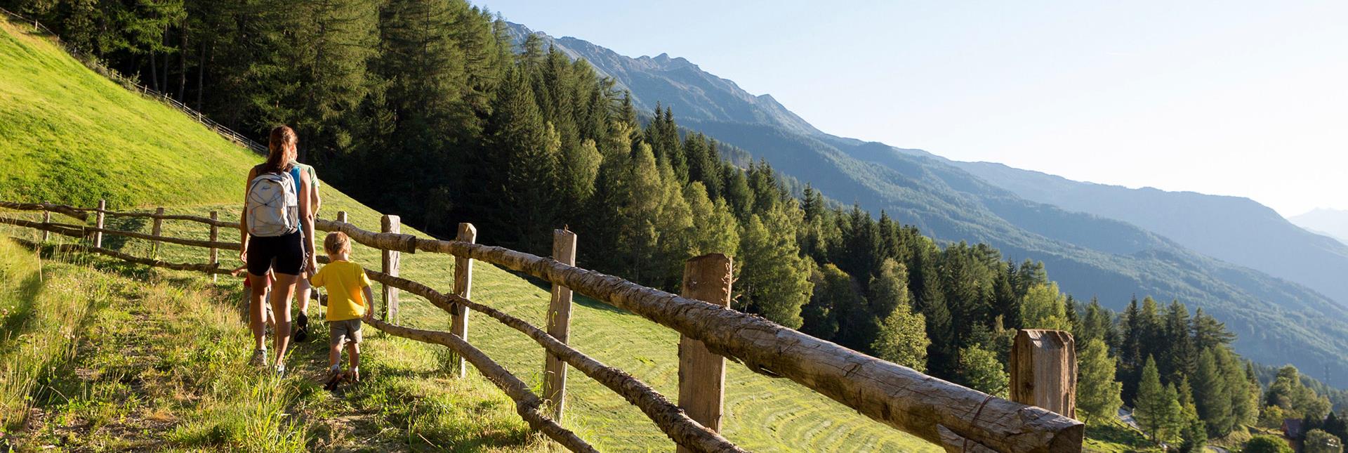 Mamma e bambino camminano su un sentiero alpino tra boschi e montagne al tramonto.