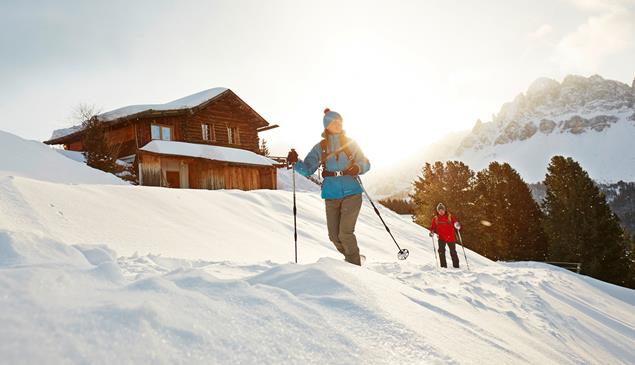 Two people snowshoe hiking past a wooden cabin and snowy mountains in the background.