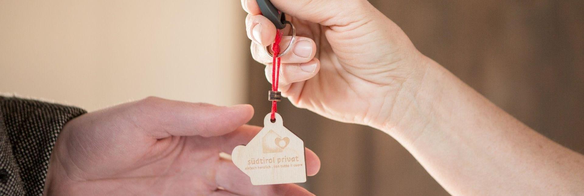 Schlüsselübergabe mit Holzanhänger „südtirol privat“ zwischen zwei Händen in Nahaufnahme.