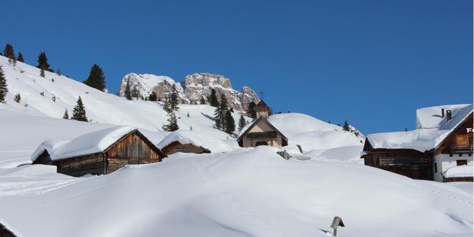 Snow-covered alpine village with wooden huts and chapel, surrounded by mountains under clear skies.
