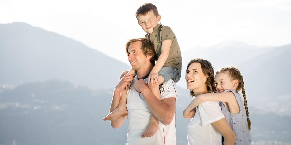 Eine fröhliche Familie mit zwei Kindern genießt den Ausblick auf die Berge – die Eltern tragen die Kinder auf den Schultern.