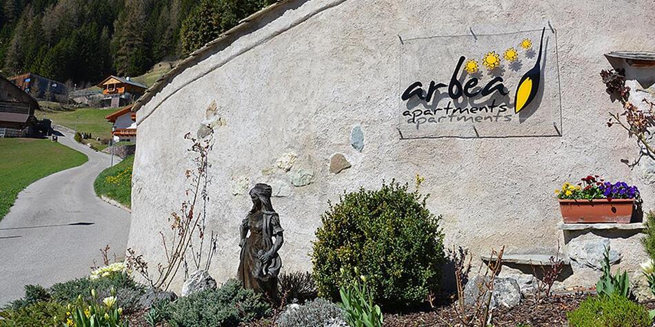 Vista di un giardino con una piccola statua di una donna in costume tradizionale davanti a un muro con l'insegna Arbea Apartments. Sullo sfondo una strada stretta, prati verdi e case in legno in un paesaggio alpino.