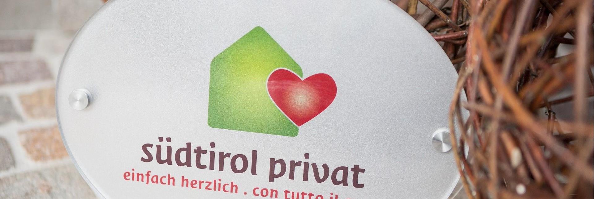 Eine Plakette mit dem Logo der Südtiroler Privatvermieter und der Aufschrift südtirol privat. einfach herzlich. con tutto il cuore