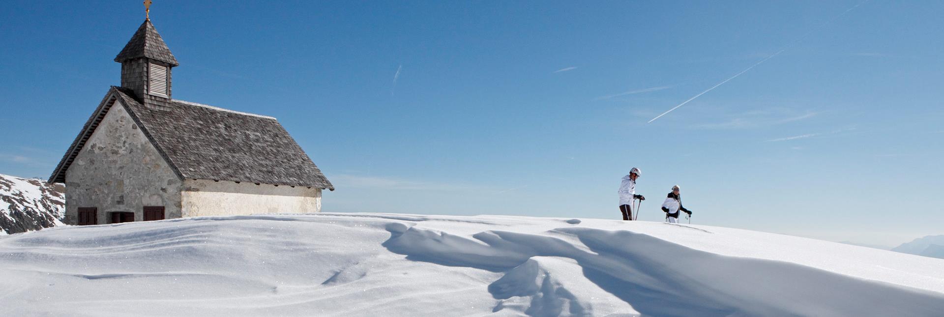 Two ski mountaineers near a chapel in a snowy winter landscape under a blue sky.