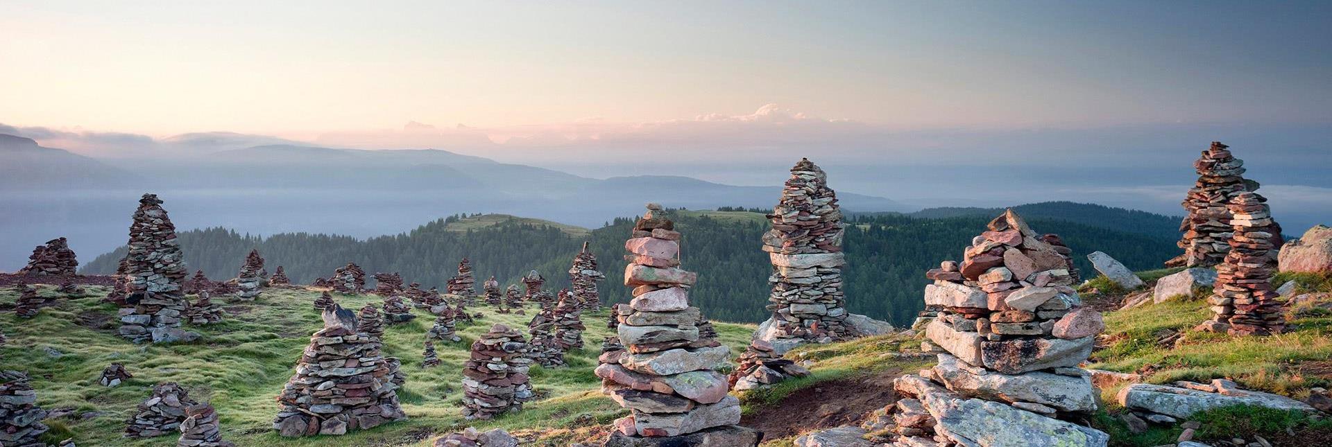 Steinmännchen auf einer Almwiese bei Sonnenaufgang mit Blick auf Hügel und Berge im Hintergrund.