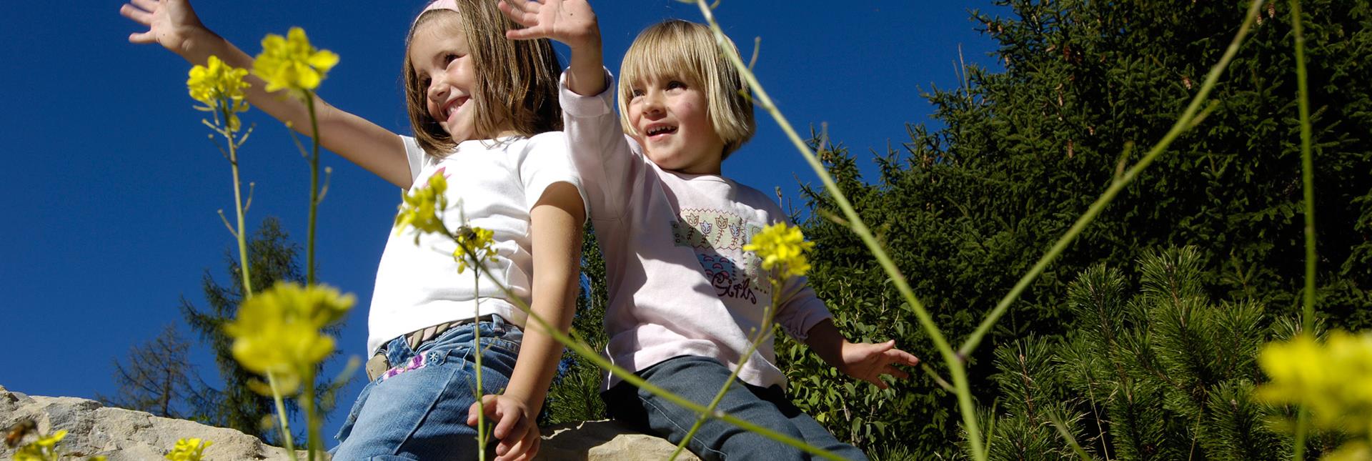 Zwei Kinder sitzen lachend auf einem Felsen und winken in die Kamera, umgeben von gelben Blumen.