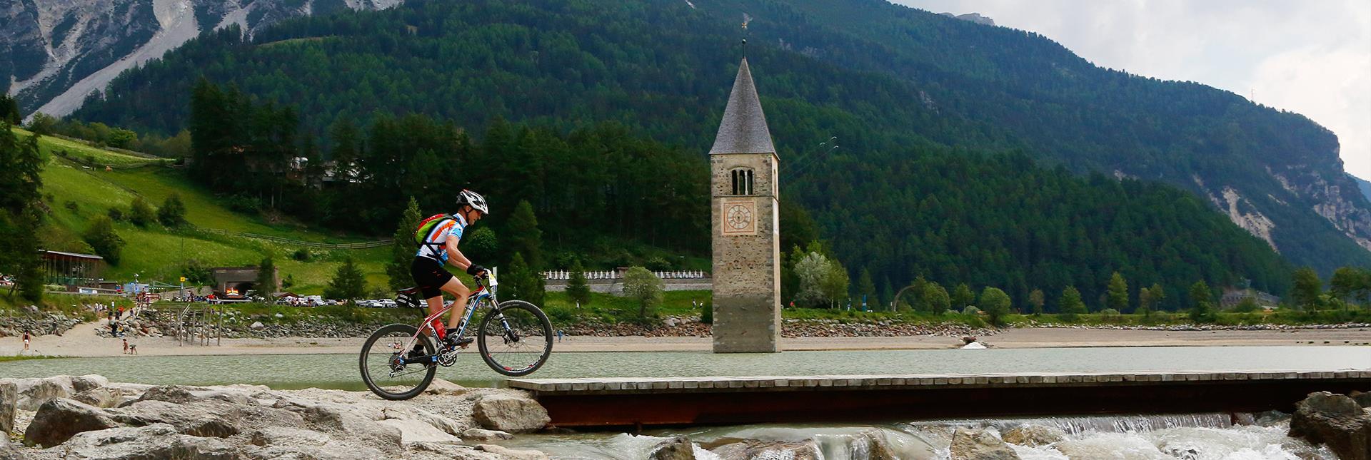 Ein Radfahrer fährt an dem versunkenen Kirchturm im Reschensee vorbei